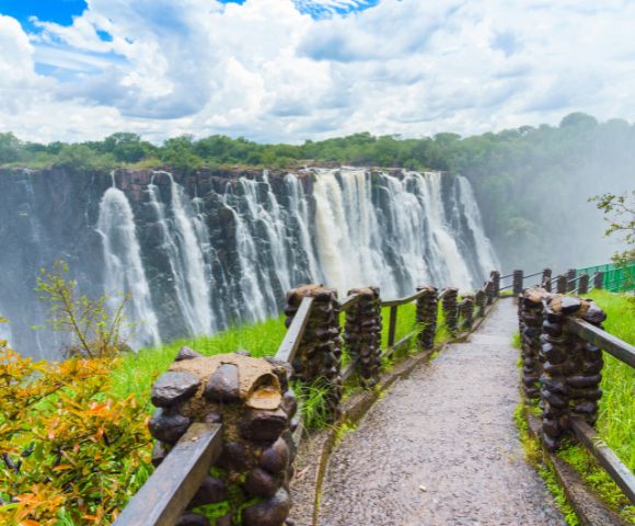 Pathway leading to a majestic waterfall with lush greenery on the sides, under a cloudy sky. The scene is vibrant and awe-inspiring.