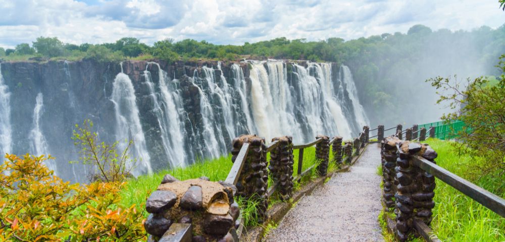 Pathway leading to a majestic waterfall with lush greenery on the sides, under a cloudy sky. The scene is vibrant and awe-inspiring.