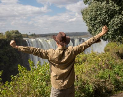 A person in a hat, arms raised in joy, faces a majestic waterfall surrounded by lush greenery under a partly cloudy sky, conveying awe and freedom.