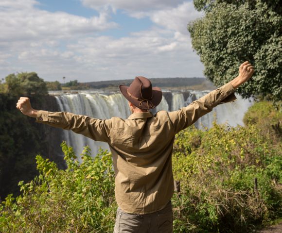 A person in a hat, arms raised in joy, faces a majestic waterfall surrounded by lush greenery under a partly cloudy sky, conveying awe and freedom.