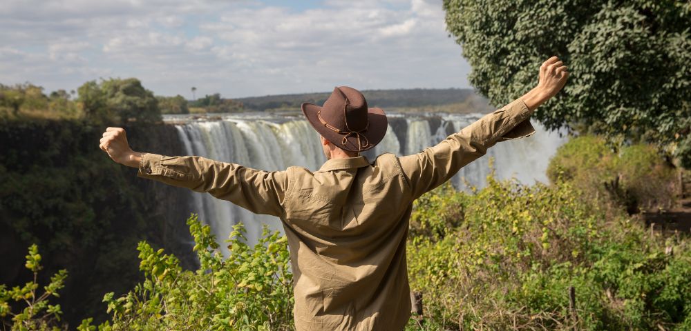 A person in a hat, arms raised in joy, faces a majestic waterfall surrounded by lush greenery under a partly cloudy sky, conveying awe and freedom.