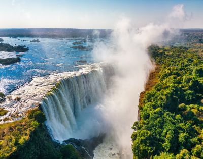 Aerial view of a majestic waterfall cascading into a gorge, surrounded by lush green forest under a clear blue sky, with mist rising dramatically.
