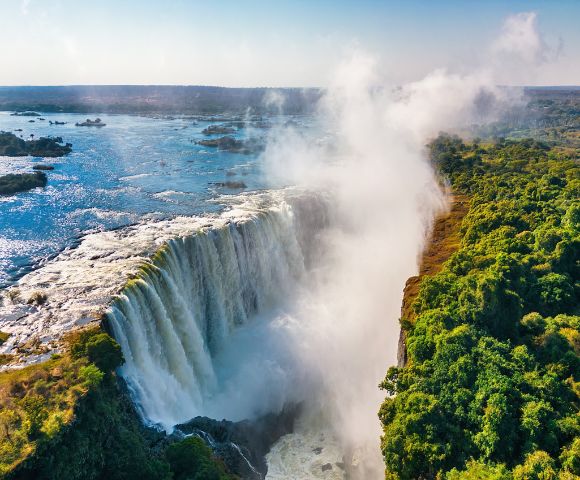 Aerial view of a majestic waterfall cascading into a gorge, surrounded by lush green forest under a clear blue sky, with mist rising dramatically.