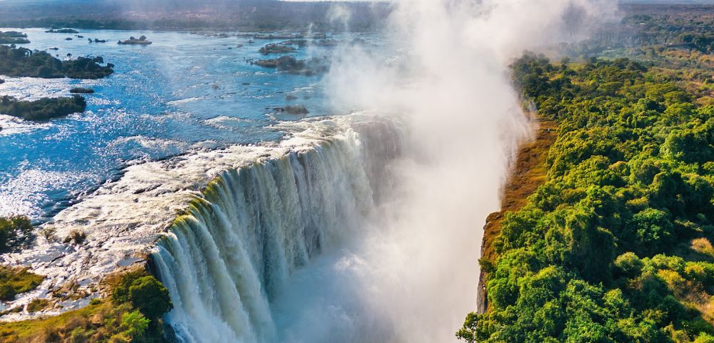 Aerial view of a majestic waterfall cascading into a gorge, surrounded by lush green forest under a clear blue sky, with mist rising dramatically.