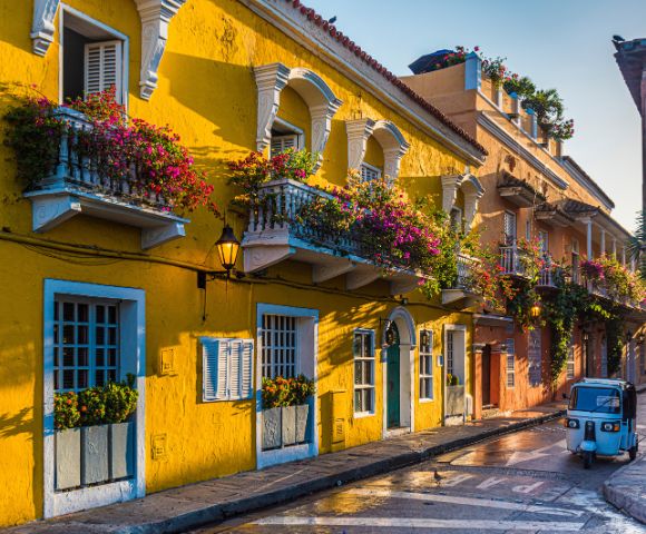 Colorful street scene with vibrant yellow buildings adorned with flowers on balconies. A small blue vehicle adds charm in the golden light of sunset.