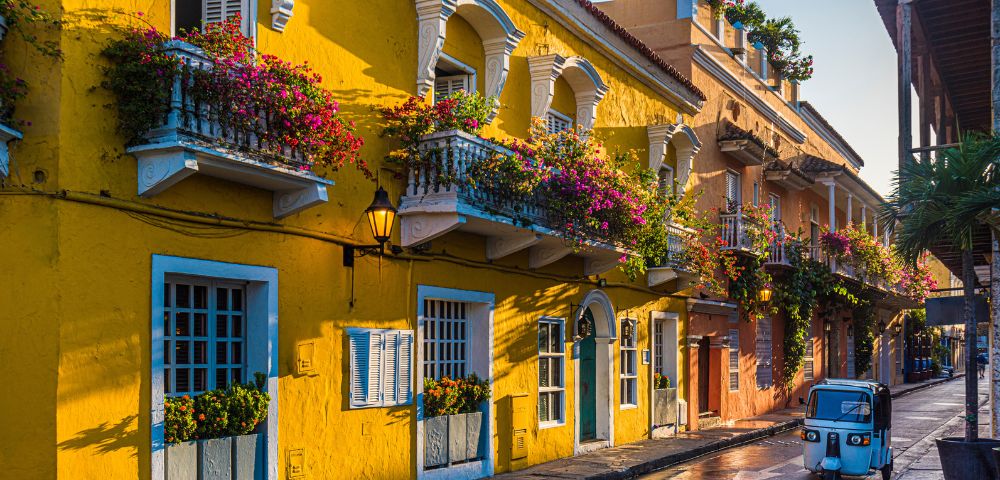 Colorful street scene with vibrant yellow buildings adorned with flowers on balconies. A small blue vehicle adds charm in the golden light of sunset.
