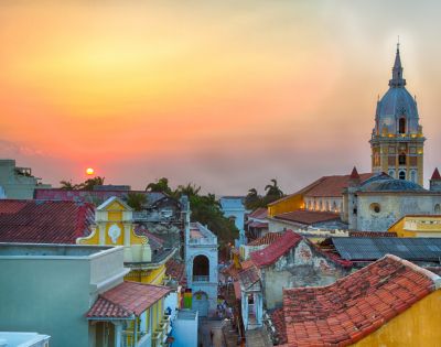 Sunset over a vibrant cityscape featuring colorful colonial buildings and a prominent church tower. The sky is warm, evoking a peaceful ambiance.