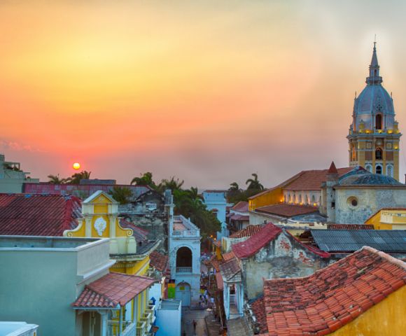 Sunset over a vibrant cityscape featuring colorful colonial buildings and a prominent church tower. The sky is warm, evoking a peaceful ambiance.