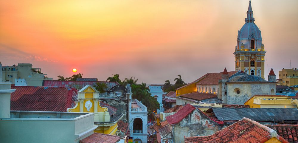 Sunset over a vibrant cityscape featuring colorful colonial buildings and a prominent church tower. The sky is warm, evoking a peaceful ambiance.