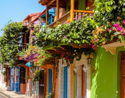 Colorful street with vibrant buildings, lush green balconies adorned with pink flowers. Bright, sunny day evokes a cheerful, lively atmosphere.