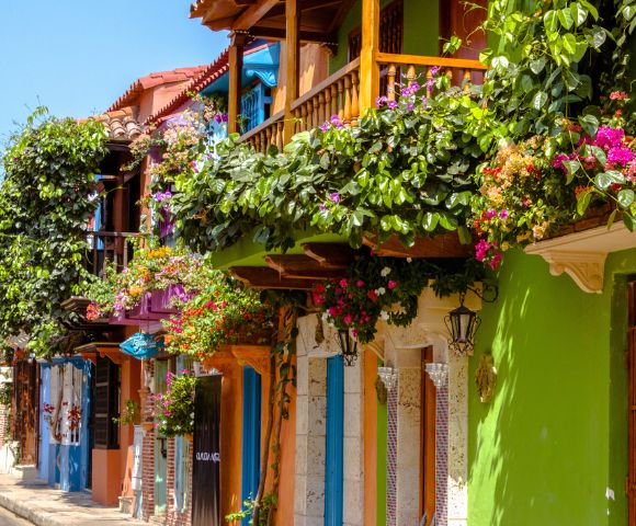 Colorful street with vibrant buildings, lush green balconies adorned with pink flowers. Bright, sunny day evokes a cheerful, lively atmosphere.