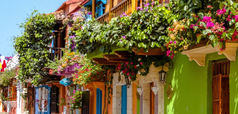 Colorful street with vibrant buildings, lush green balconies adorned with pink flowers. Bright, sunny day evokes a cheerful, lively atmosphere.