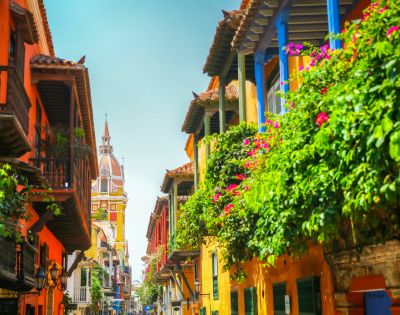 A vibrant street scene in Cartagena with colorful buildings, ornate balconies, lush green plants, and a distant cathedral spire under a clear blue sky.
