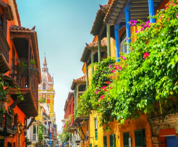 A vibrant street scene in Cartagena with colorful buildings, ornate balconies, lush green plants, and a distant cathedral spire under a clear blue sky.