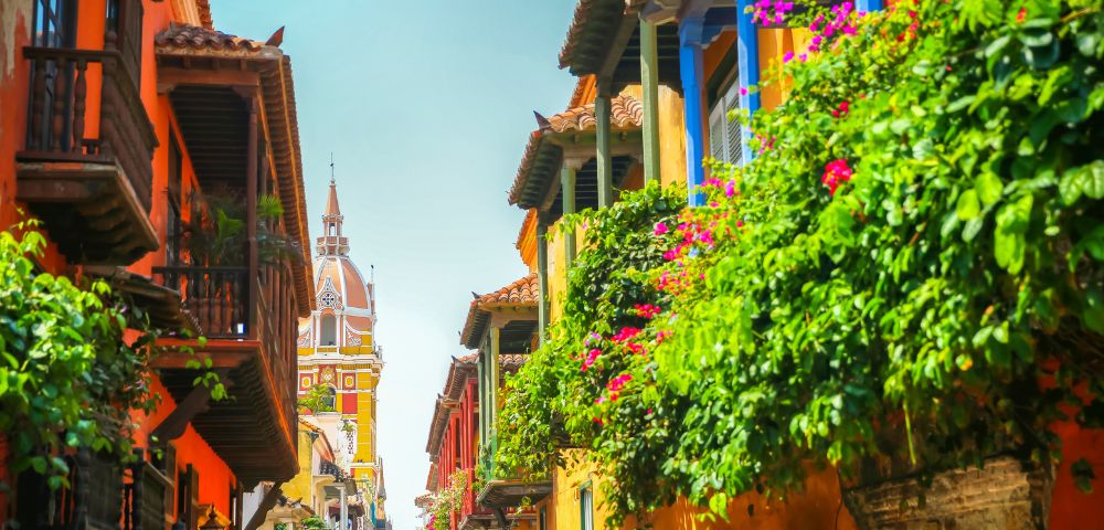 A vibrant street scene in Cartagena with colorful buildings, ornate balconies, lush green plants, and a distant cathedral spire under a clear blue sky.