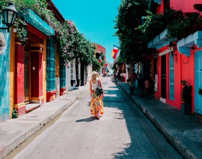A person in a floral dress walks down a colorful street with vibrant red, blue, and orange buildings. Sunlit, lively atmosphere with potted plants.