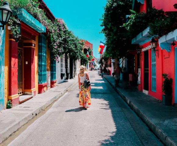 A person in a floral dress walks down a colorful street with vibrant red, blue, and orange buildings. Sunlit, lively atmosphere with potted plants.
