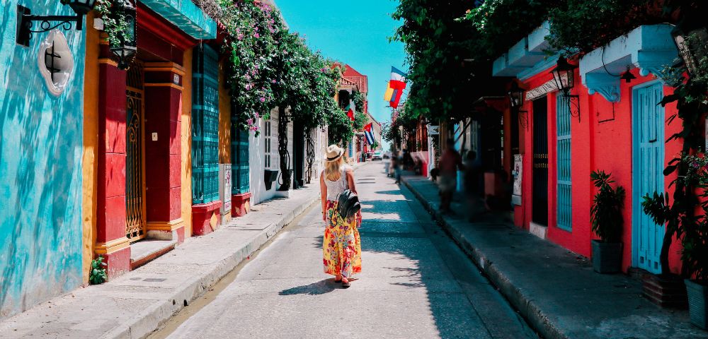 A person in a floral dress walks down a colorful street with vibrant red, blue, and orange buildings. Sunlit, lively atmosphere with potted plants.