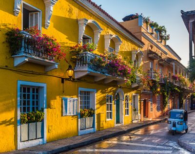 Colorful street scene with vibrant yellow buildings adorned with flowers on balconies. A small blue vehicle adds charm in the golden light of sunset.