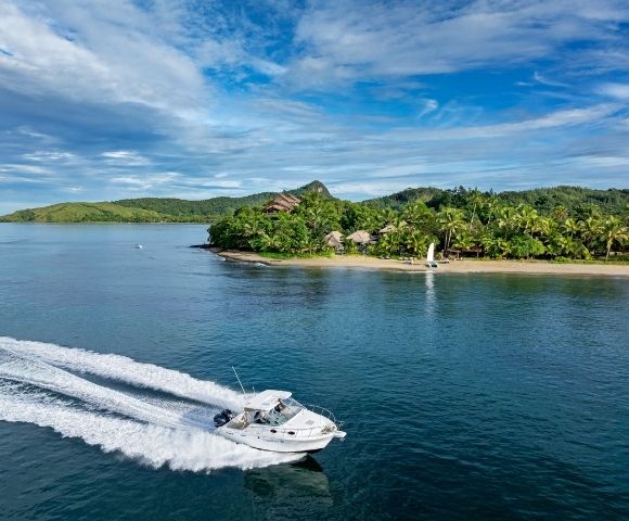 A speedboat creates white waves against clear blue water near a tropical island with lush greenery and a sandy beach on a sunny day.