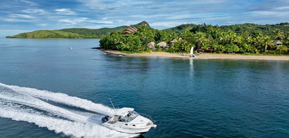 A speedboat creates white waves against clear blue water near a tropical island with lush greenery and a sandy beach on a sunny day.
