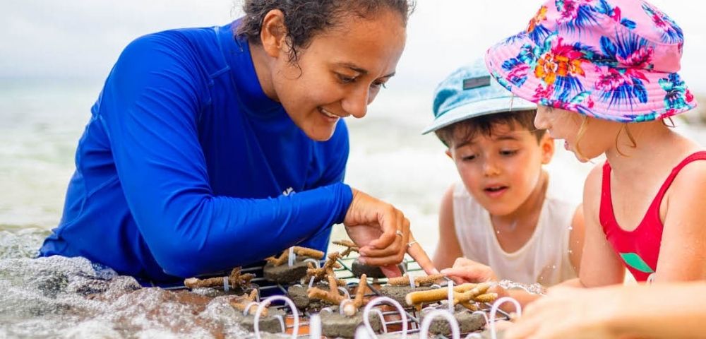 A woman and two children play with a wooden toy structure on a sunny beach. The children wear colorful hats, looking engaged and curious.