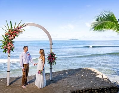 A couple stands under a floral arch on a sandy beach, gazing at each other. The ocean and distant island are in the background, creating a serene and romantic atmosphere.