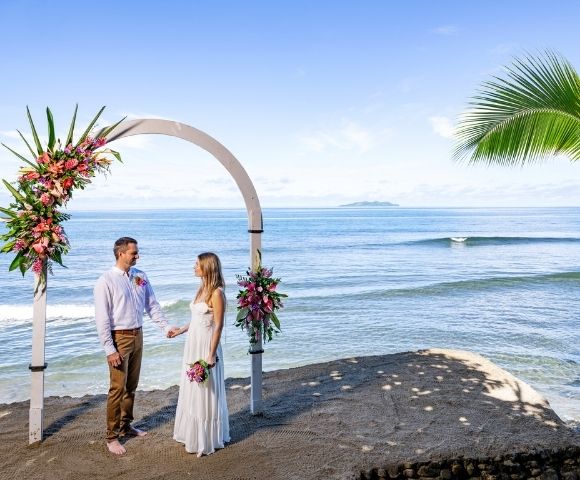 A couple stands under a floral arch on a sandy beach, gazing at each other. The ocean and distant island are in the background, creating a serene and romantic atmosphere.