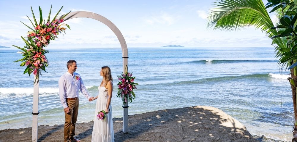 A couple stands under a floral arch on a sandy beach, gazing at each other. The ocean and distant island are in the background, creating a serene and romantic atmosphere.