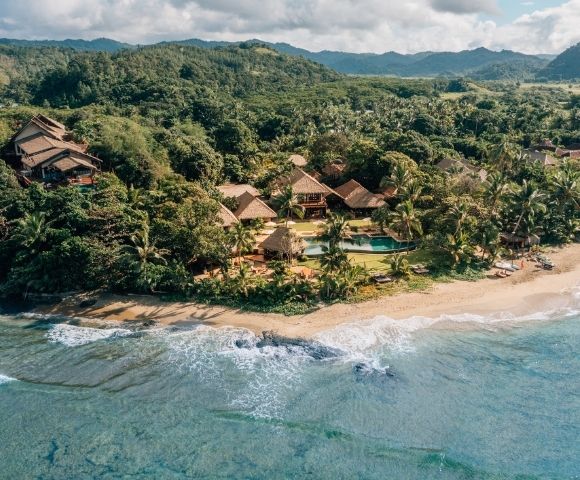 Aerial view of a lush tropical resort with wooden villas nestled among dense greenery, bordering a serene sandy beach with gentle ocean waves.