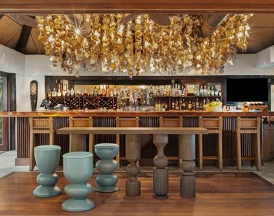 Elegant bar interior with a warm ambiance; wooden stools and tables. Overhead, a decorative ceiling adorned with golden hanging ornaments. Bottles line the back shelves.