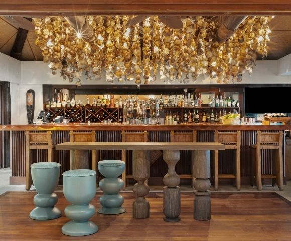Elegant bar interior with a warm ambiance; wooden stools and tables. Overhead, a decorative ceiling adorned with golden hanging ornaments. Bottles line the back shelves.