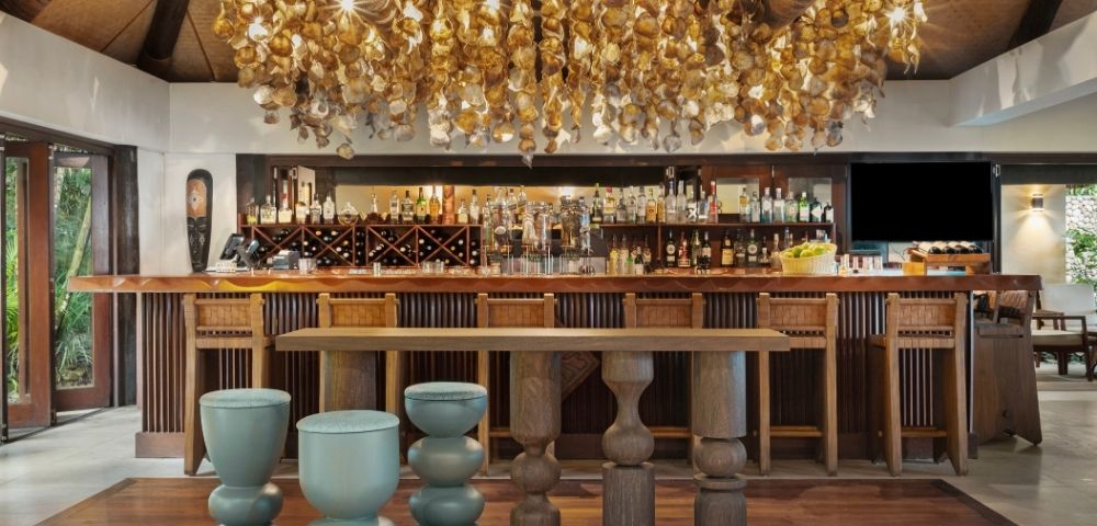 Elegant bar interior with a warm ambiance; wooden stools and tables. Overhead, a decorative ceiling adorned with golden hanging ornaments. Bottles line the back shelves.