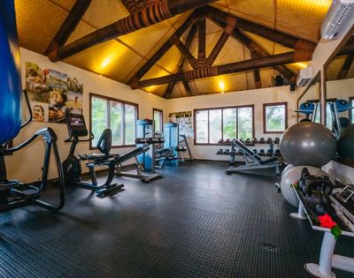 A well-lit gym features exercise bikes, weight benches, and stability balls under a wooden beam ceiling. Motivational posters adorn the walls.