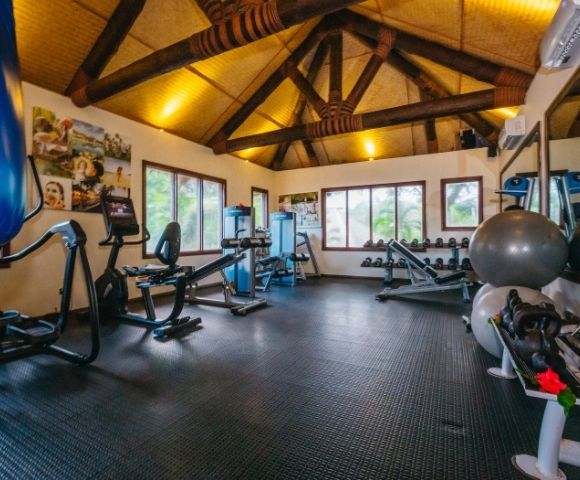 A well-lit gym features exercise bikes, weight benches, and stability balls under a wooden beam ceiling. Motivational posters adorn the walls.