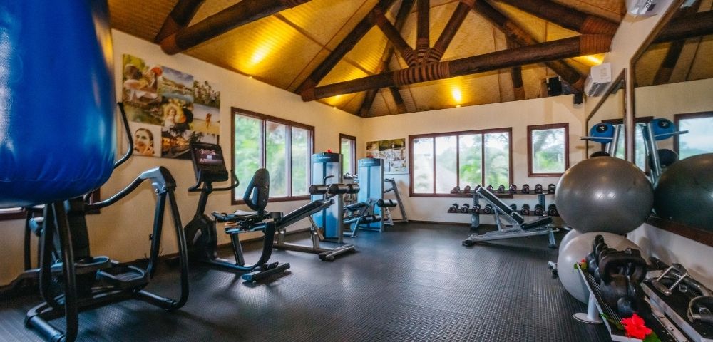 A well-lit gym features exercise bikes, weight benches, and stability balls under a wooden beam ceiling. Motivational posters adorn the walls.