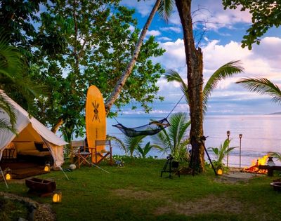A tranquil beachside campsite with a canvas tent, surfboard, hammock, and campfire. Tropical trees and ocean views create a serene atmosphere.