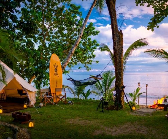 A tranquil beachside campsite with a canvas tent, surfboard, hammock, and campfire. Tropical trees and ocean views create a serene atmosphere.