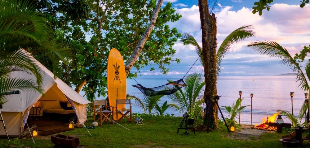 A tranquil beachside campsite with a canvas tent, surfboard, hammock, and campfire. Tropical trees and ocean views create a serene atmosphere.