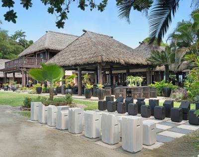 Traditional Polynesian-style thatched-roof buildings surrounded by lush greenery and tropical plants. Decor includes white and black stone stools.