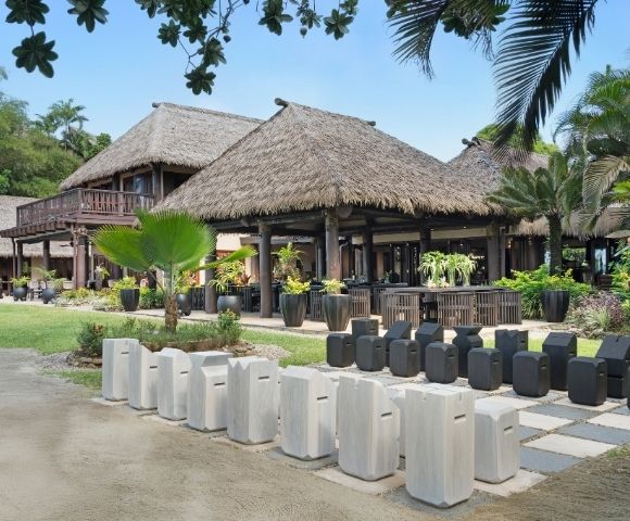 Traditional Polynesian-style thatched-roof buildings surrounded by lush greenery and tropical plants. Decor includes white and black stone stools.