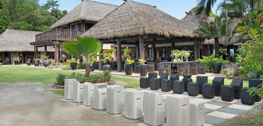 Traditional Polynesian-style thatched-roof buildings surrounded by lush greenery and tropical plants. Decor includes white and black stone stools.