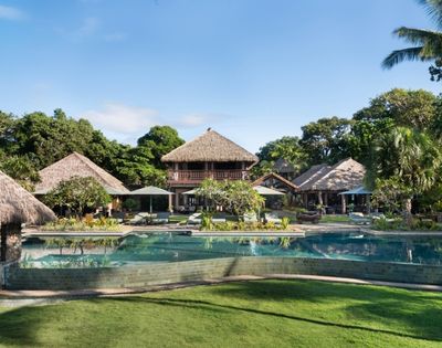 A tranquil tropical resort features thatched-roof buildings surrounded by lush greenery. A large, inviting pool and well-maintained lawn are in the foreground.