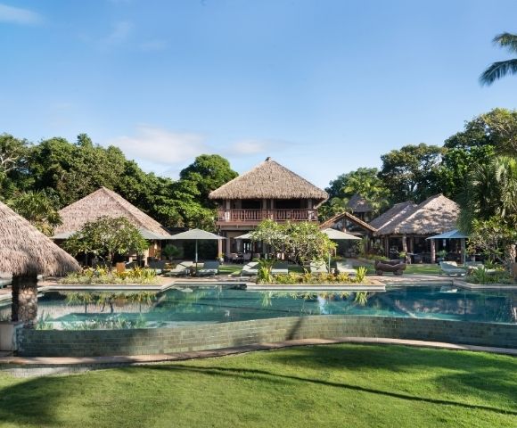 A tranquil tropical resort features thatched-roof buildings surrounded by lush greenery. A large, inviting pool and well-maintained lawn are in the foreground.