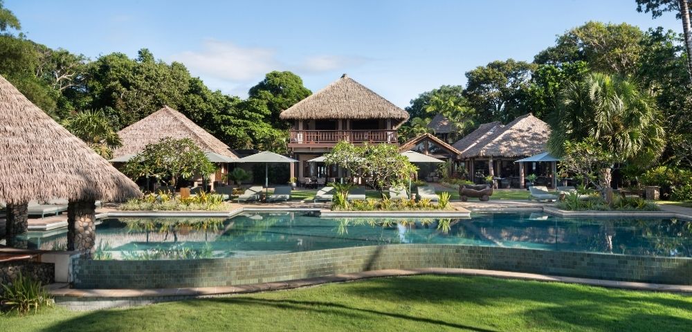A tranquil tropical resort features thatched-roof buildings surrounded by lush greenery. A large, inviting pool and well-maintained lawn are in the foreground.