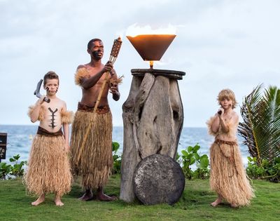 A man and two boys in grass skirts perform a traditional torch-lighting ritual by the ocean. The scene conveys cultural heritage and ceremonial respect.