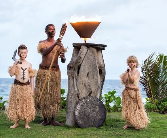 A man and two boys in grass skirts perform a traditional torch-lighting ritual by the ocean. The scene conveys cultural heritage and ceremonial respect.