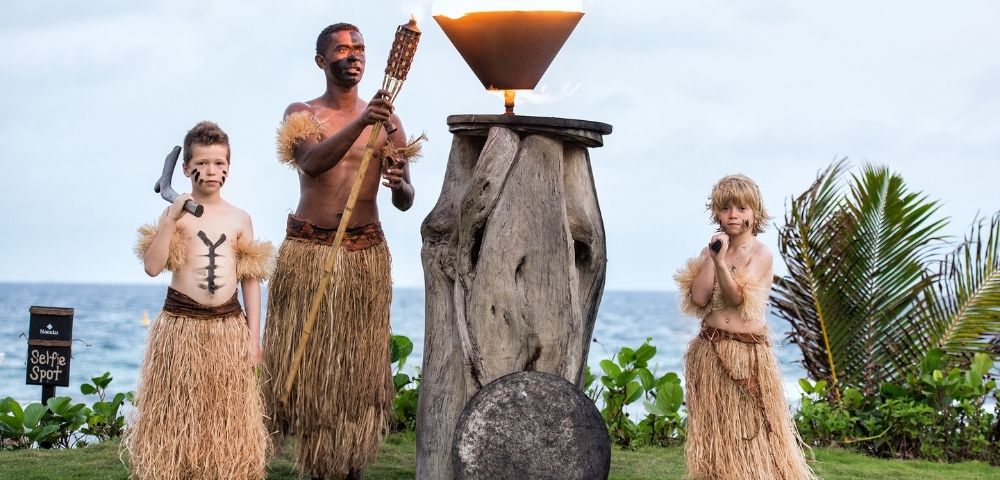 A man and two boys in grass skirts perform a traditional torch-lighting ritual by the ocean. The scene conveys cultural heritage and ceremonial respect.
