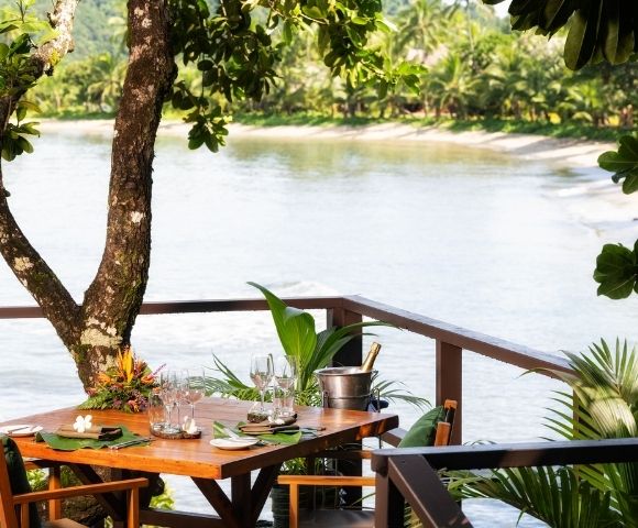 A tropical dining setup overlooks a serene, sandy beach and calm sea. A wooden table with elegant place settings, chairs, and a champagne bucket under lush greenery.