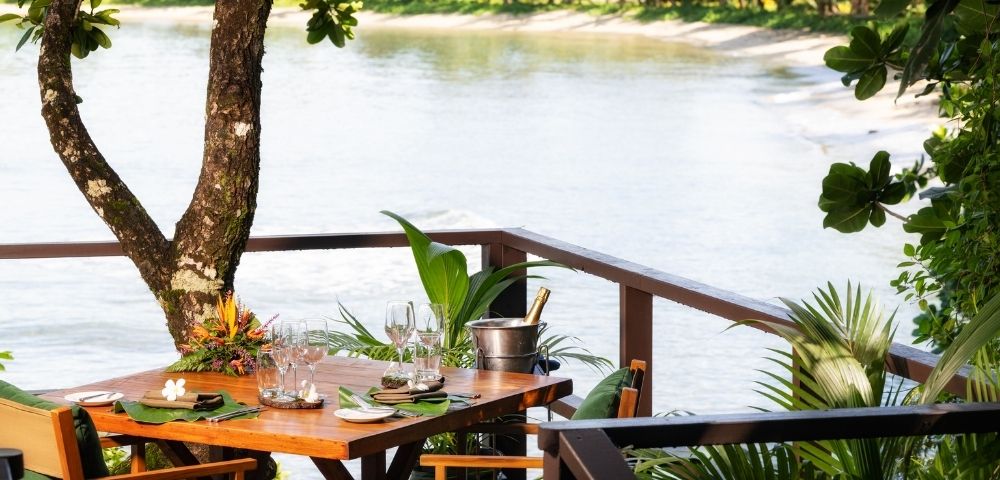 A tropical dining setup overlooks a serene, sandy beach and calm sea. A wooden table with elegant place settings, chairs, and a champagne bucket under lush greenery.